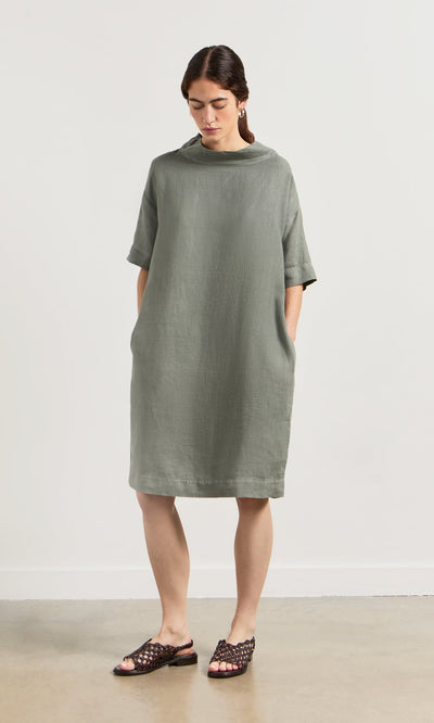 Plümo- Woman in the sage green Sims Dress—a loose linen shift with crop sleeves and pockets—pairing woven sandals.