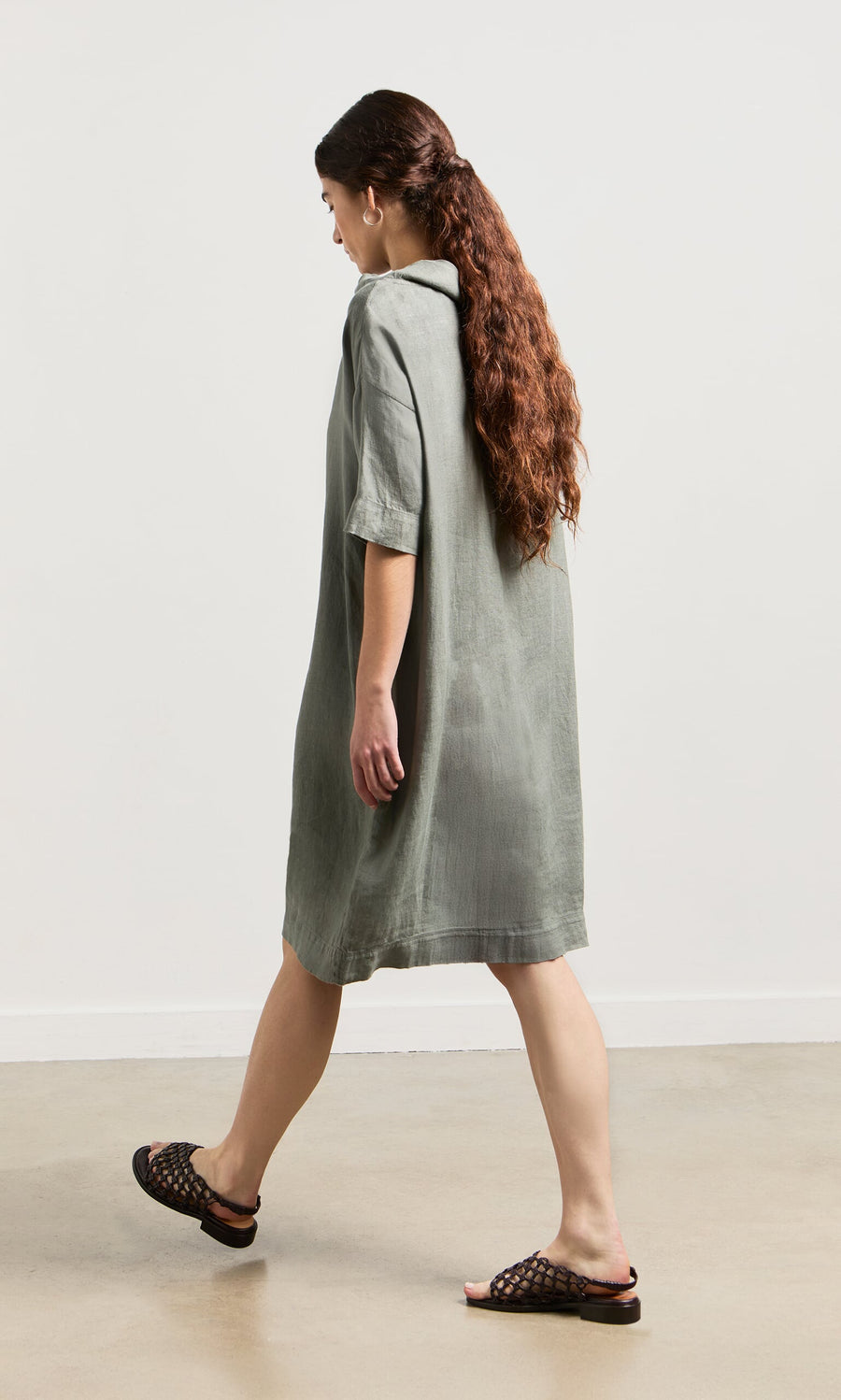 Plümo- A woman with long brown hair in the Sims Dress and black sandals walks indoors on a light-colored floor.