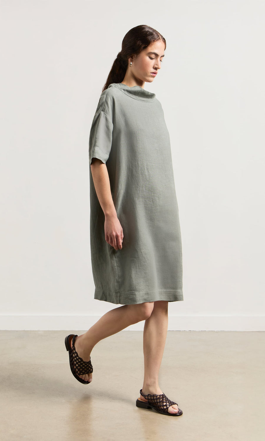 Plümo- Woman wearing the Sims Dress, a loose gray-green linen shift with crop sleeves, and black sandals indoors by a white wall.