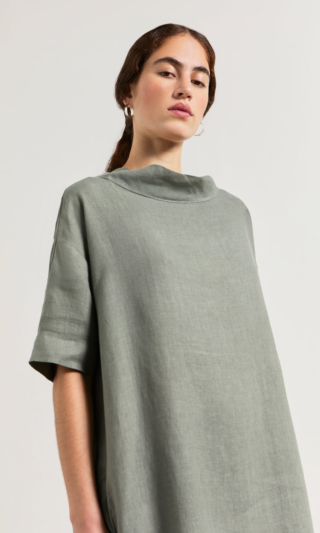 Plümo- Woman in the loose sage green Sims Dress and hoop earrings, posing against a plain background.