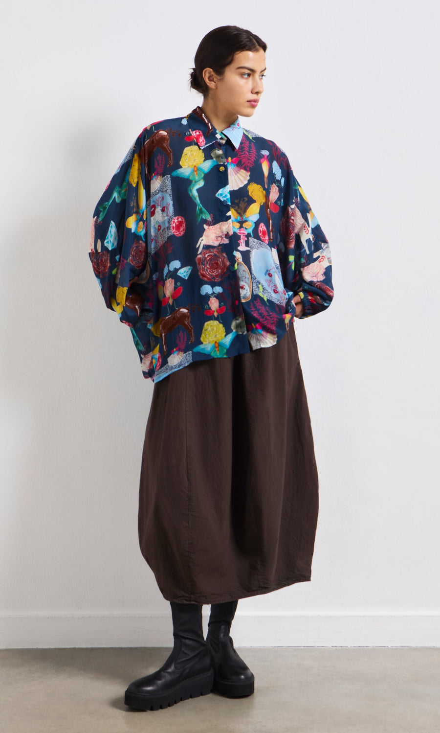 Plümo- Person wearing the Sketch Blouse, a colorful oversized shirt, with a brown wide skirt and black boots against a white wall.