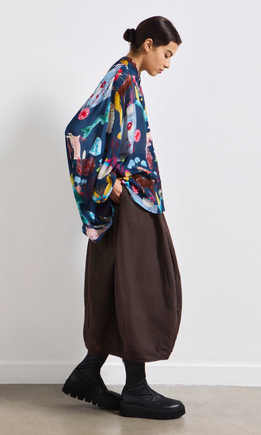 Plümo- Woman in the Sketch Blouse stands in profile against a plain white wall, styled with a brown skirt and black boots.