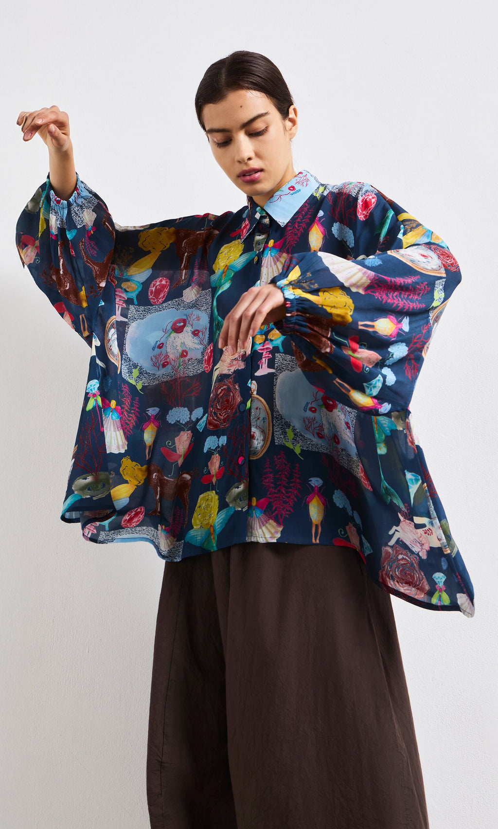 Plümo- A woman wears the Sketch Blouse—an oversized, sheer top with colorful abstract prints and wide sleeves—plus dark pants.