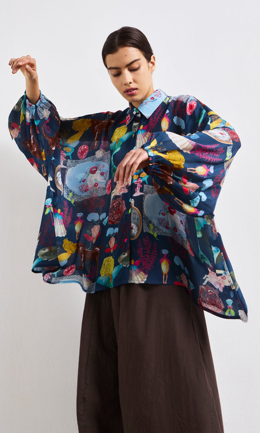 Plümo- A woman wears the Sketch Blouse—an oversized, sheer top with colorful abstract prints and wide sleeves—plus dark pants.