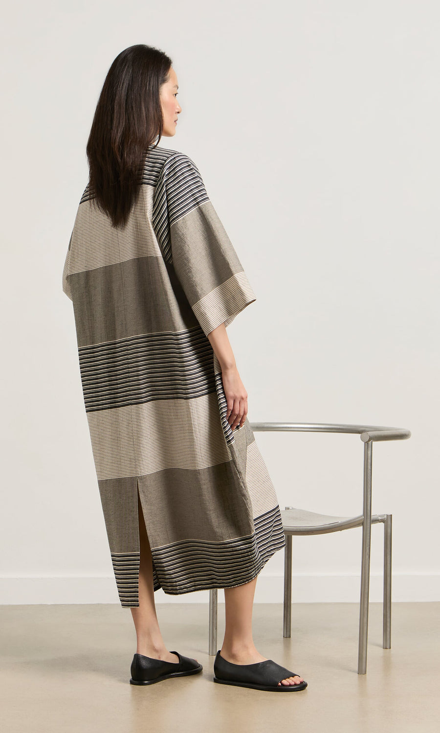 Plümo- In the Stonebar Dress and black sandals, a woman stands by a silver chair, her sculptural silhouette facing away.