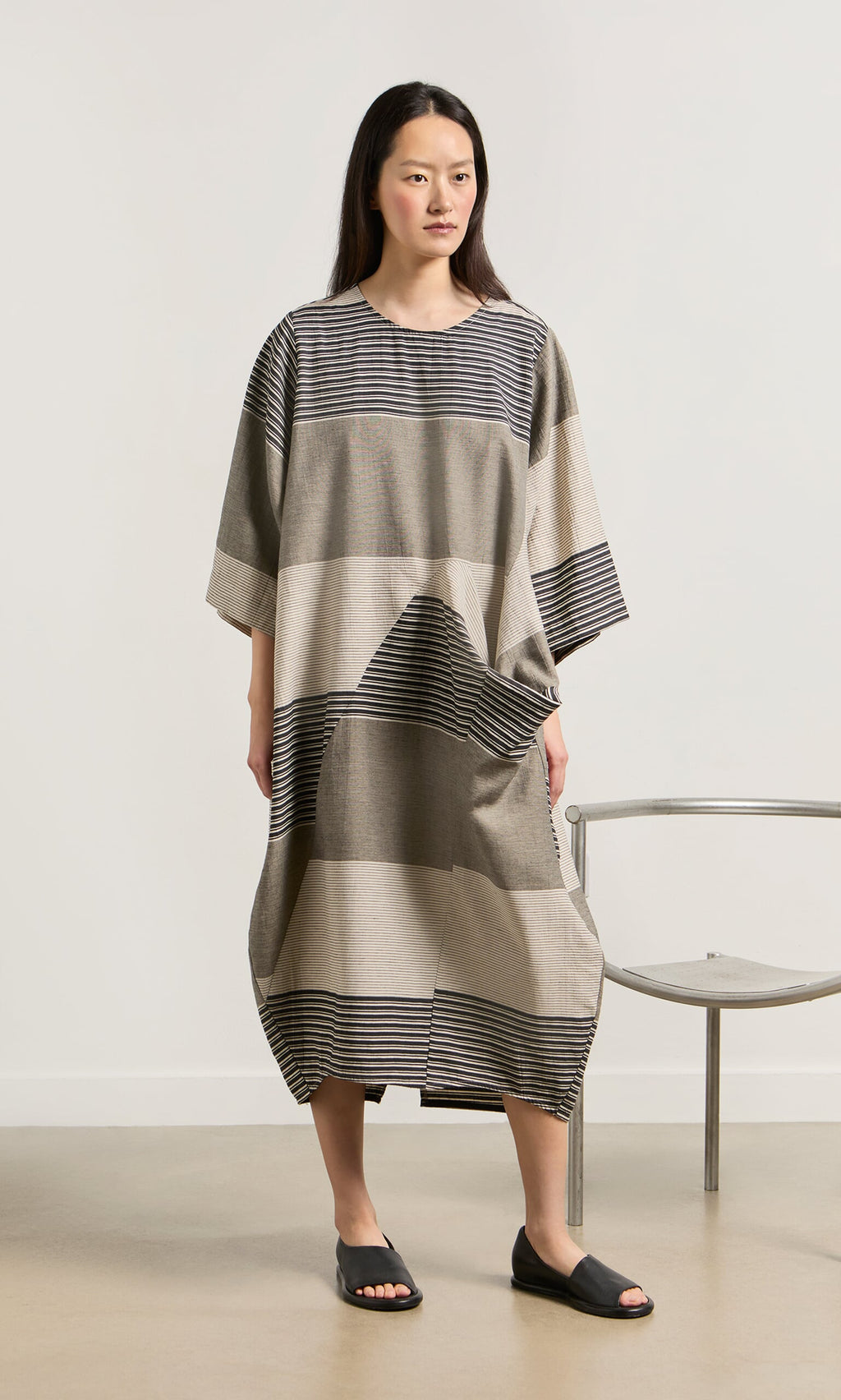 Plümo- Woman wears the Stonebar Dress in striped taupe and gray, standing indoors by a chair in black slide sandals.