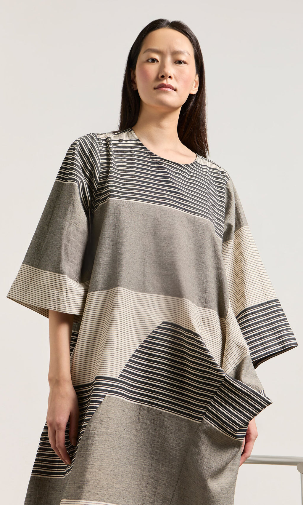 Plümo- Woman in the Stonebar Dress, a patchwork monochrome piece with wide sleeves, geometric stripes, and sculptural silhouette.