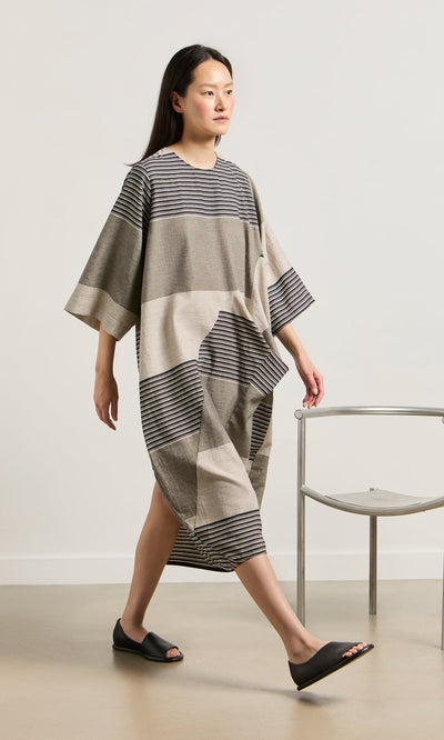 Plümo- A woman in the Stonebar Dress walks past a modern metal chair, wearing black slide sandals.
