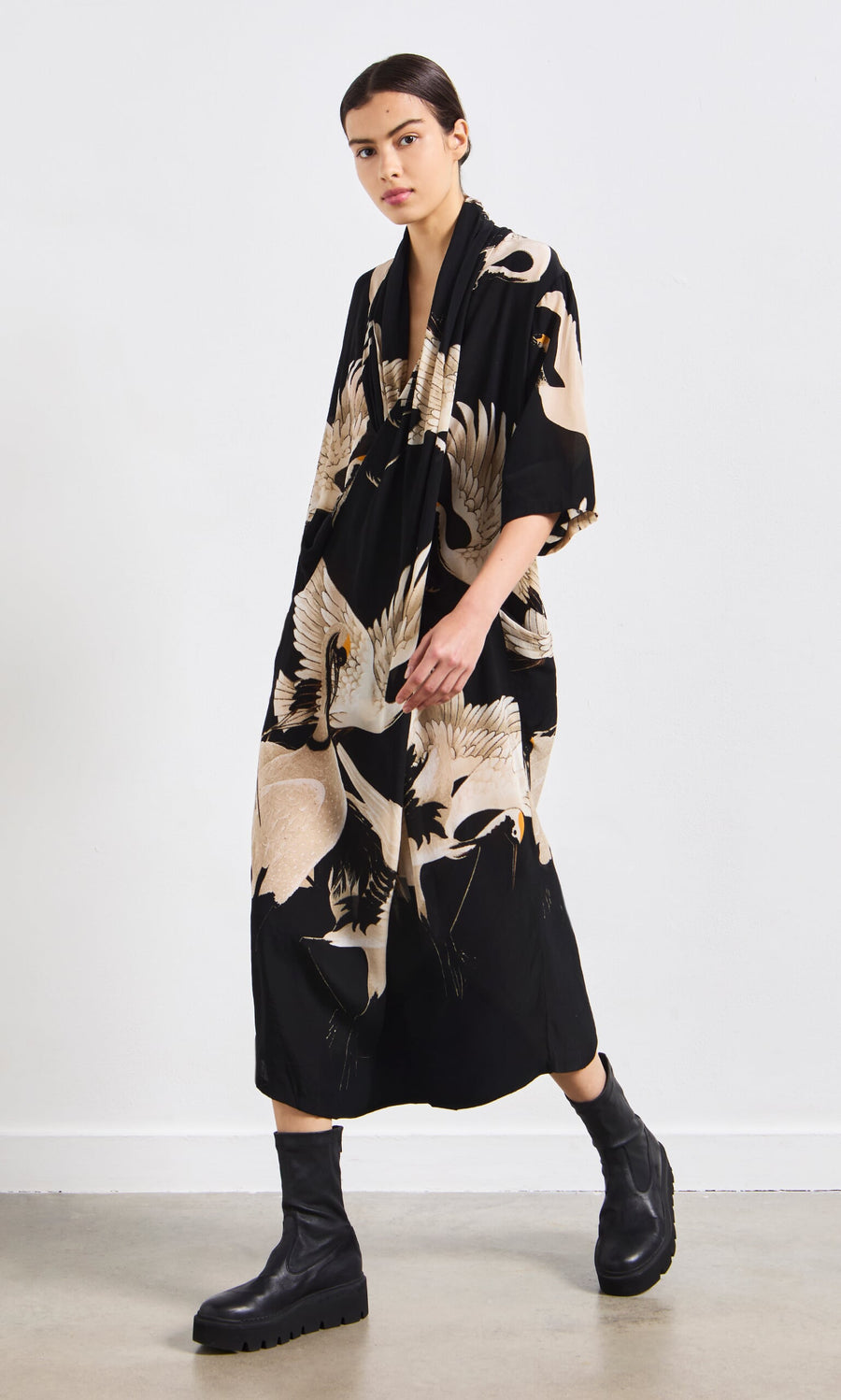 Plümo- Person wearing the Stork Twist Dress with black boots walks indoors on a polished floor.