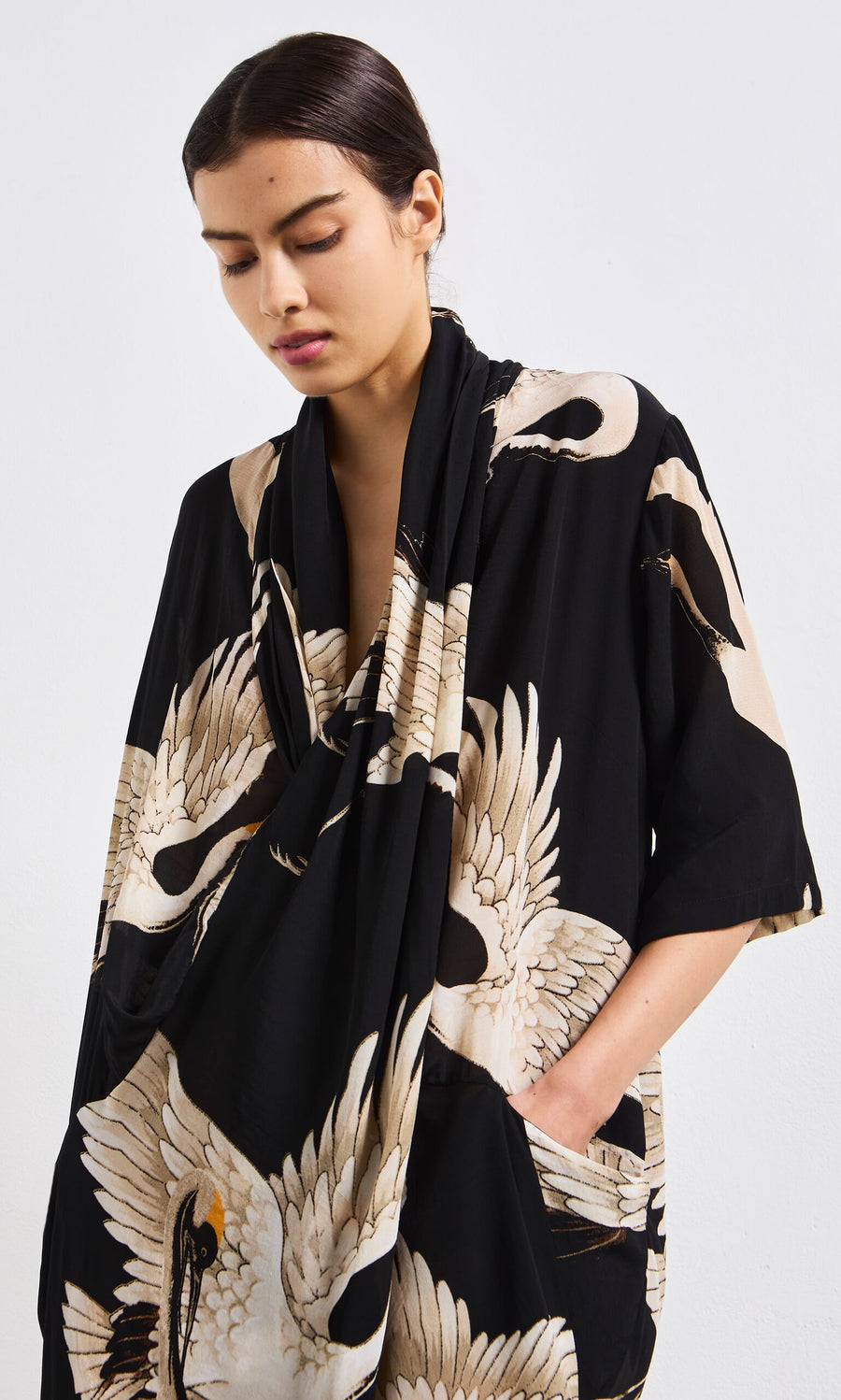 Plümo- Woman with dark hair in the Stork Twist Dress, vintage inspired with white cranes, looking down, hands in pockets.
