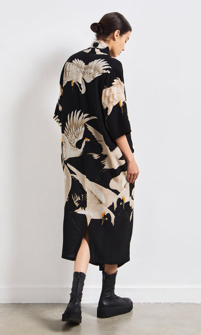 Plümo- Woman in Stork Twist Dress and black boots with a low bun, walking away on a white background.
