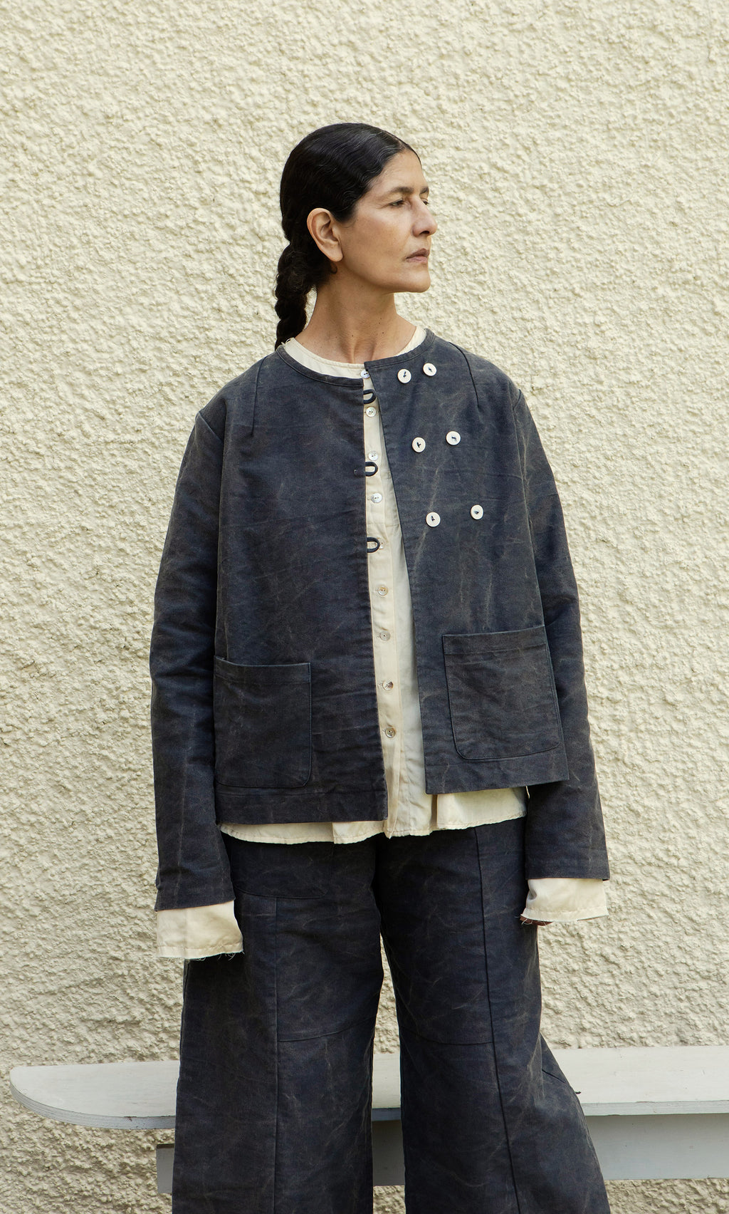 Plümo  Person in loose, layered Stour Jacket and pants stands against a textured beige wall, looking to the left.