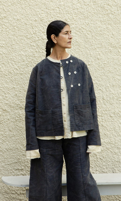 Plümo  Person in loose, layered Stour Jacket and pants stands against a textured beige wall, looking to the left.