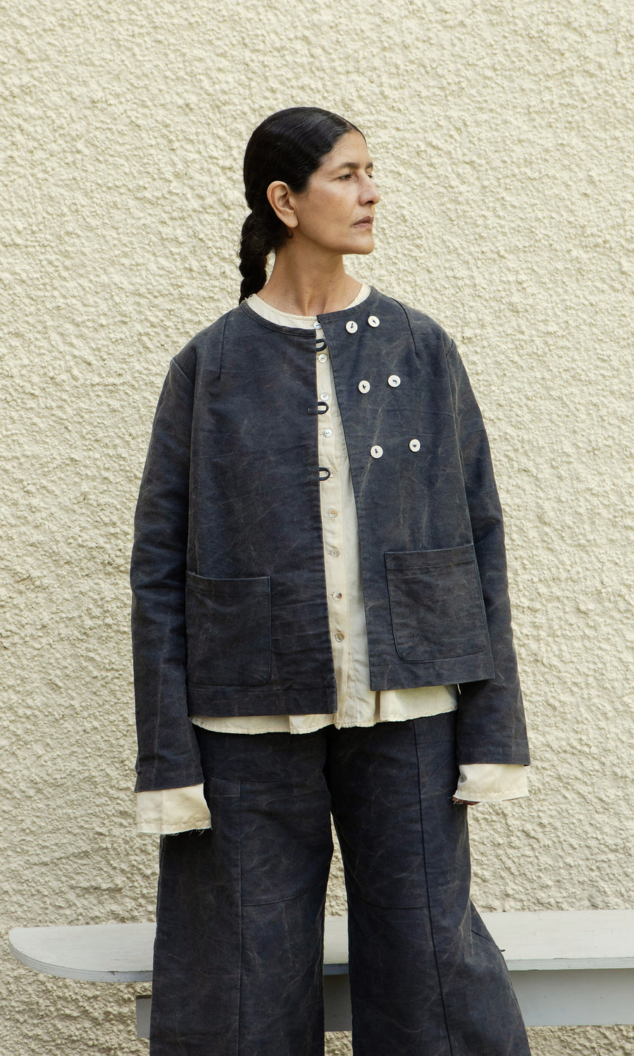 Plümo  Person in loose, layered Stour Jacket and pants stands against a textured beige wall, looking to the left.
