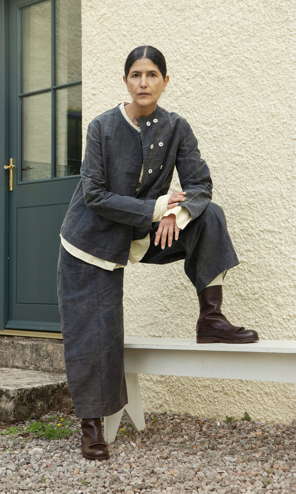 Plümo  Someone in dark, loose clothing and the Stour Jacket poses by a blue door, foot resting on the bench.