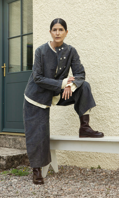 Plümo  Someone in dark, loose clothing and the Stour Jacket poses by a blue door, foot resting on the bench.