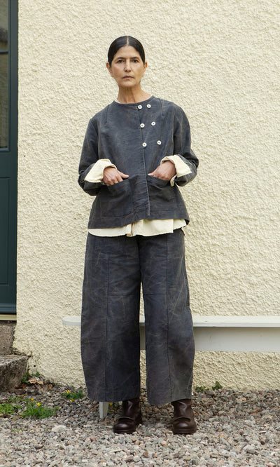Plümo  Woman stands on gravel, wearing the ethically made Stour Jacket and pants with boots in front of a wall.
