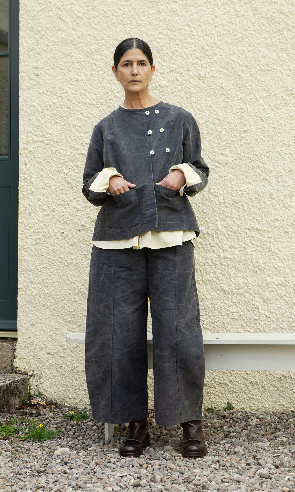 Plümo  A woman in Corfe Barrel Pants and a loose grey jacket stands on gravel before a textured cream wall.