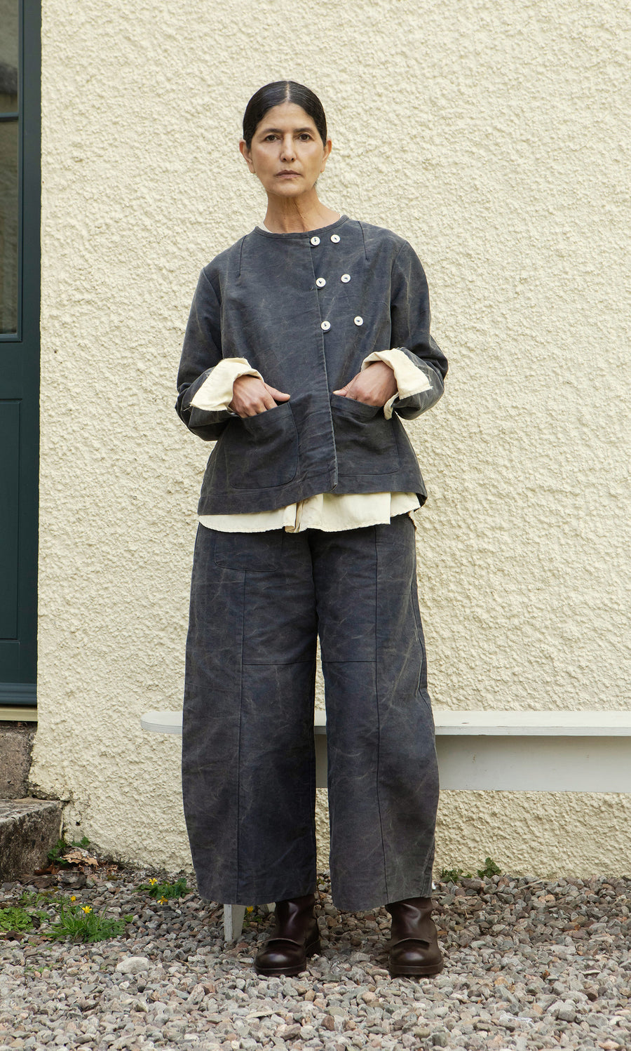 Plümo  A woman in Corfe Barrel Pants and a loose grey jacket stands on gravel before a textured cream wall.