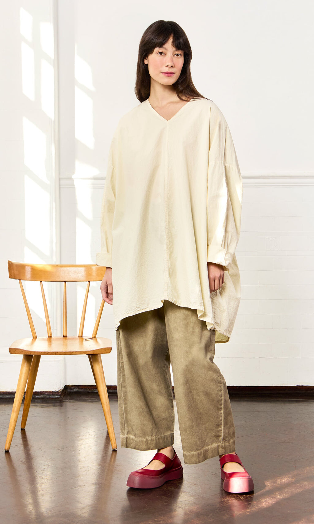 Plümo- Woman in a Stroh Blouse and khaki pants stands by a wooden chair, wearing red platform shoes in a bright room.