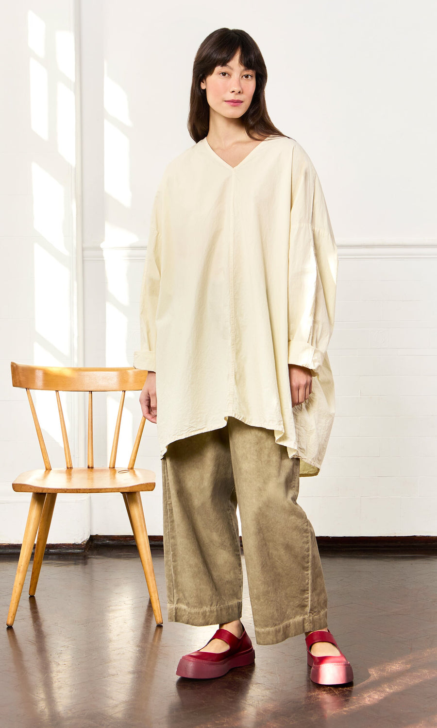 Plümo- Woman in a Stroh Blouse and khaki pants stands by a wooden chair, wearing red platform shoes in a bright room.