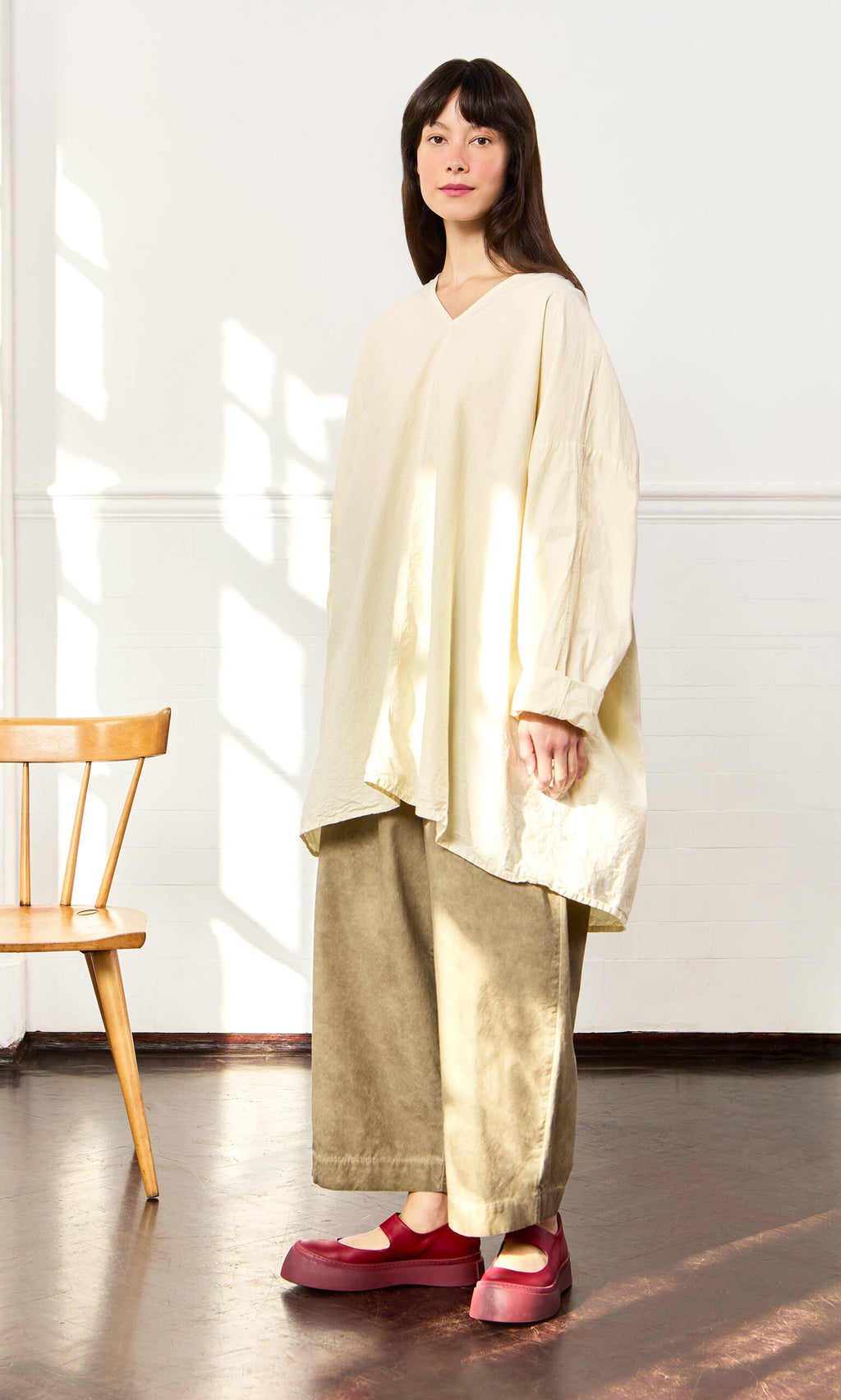 Plümo- Woman in a Stroh Blouse, wide khaki pants, and red shoes stands indoors beside a wooden chair.