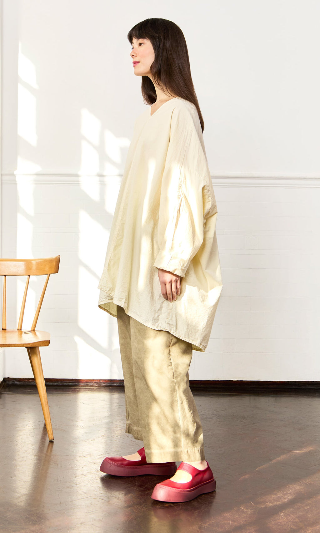 Plümo- Woman in a Stroh Blouse, beige pants, and red shoes stands indoors by a wooden chair in sunlight.