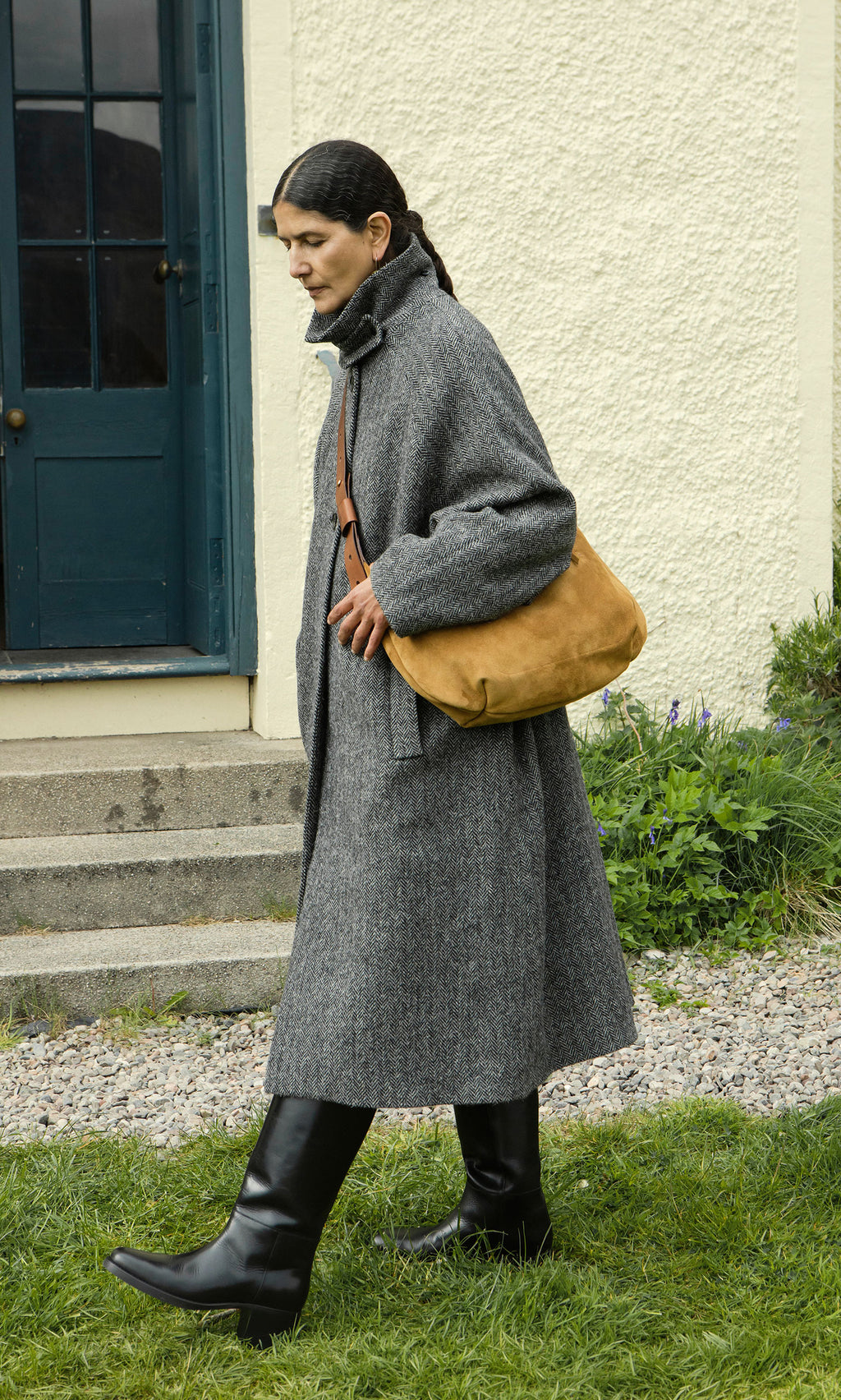 Plümo  Woman in a gray overcoat and black boots carries the Kiel Suede Bag outside near a blue door.