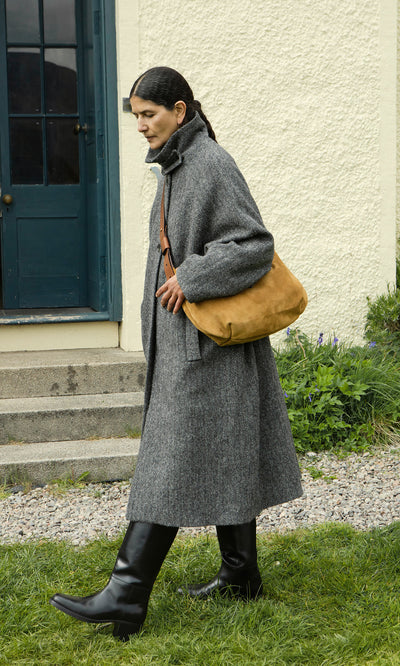 Plümo  Woman in a gray overcoat and black boots carries the Kiel Suede Bag outside near a blue door.