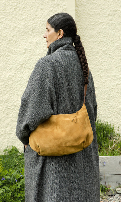 Plümo  Woman in a textured gray coat with a long braid and Kiel Suede Bag stands by greenery.