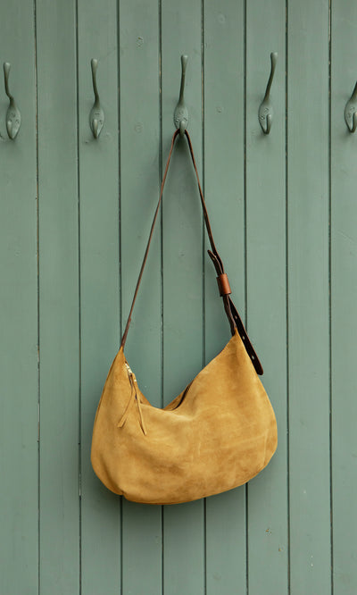 Plümo  The Kiel Suede Bag in tan Italian leather hangs on a green wooden door with four decorative hooks.