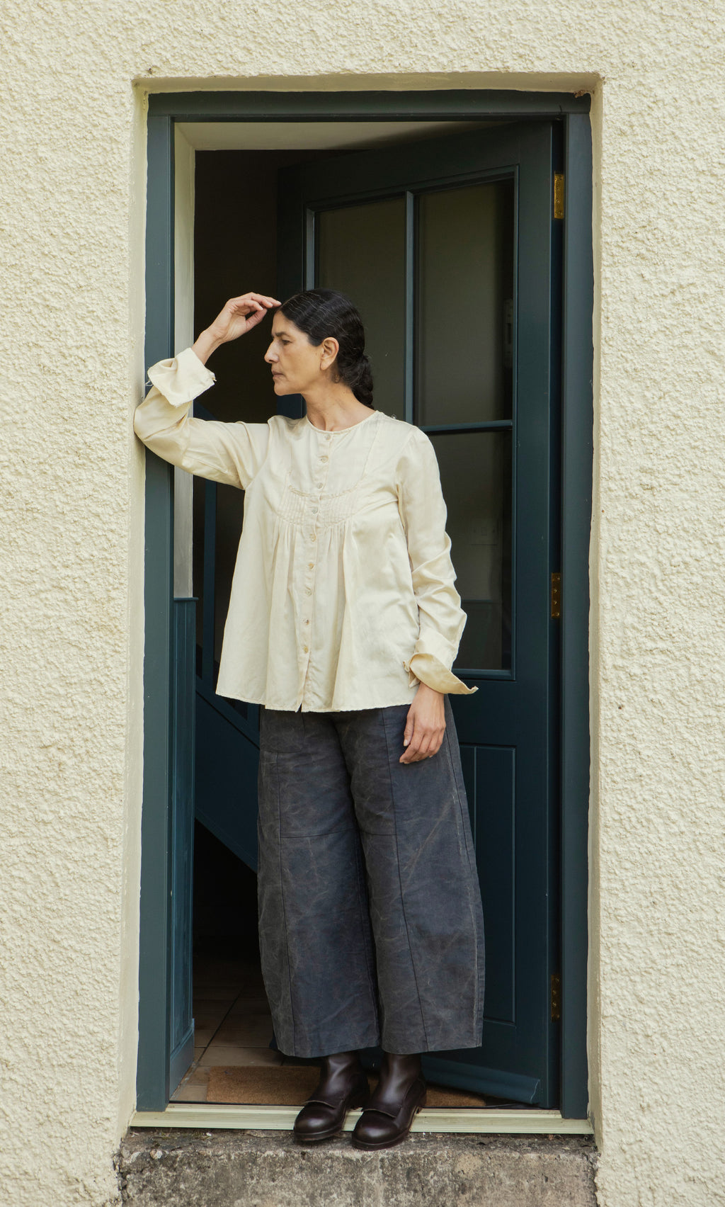 Plümo  Woman in a cream blouse and Corfe Barrel Pants stands thoughtfully in a doorway, looking to her left.