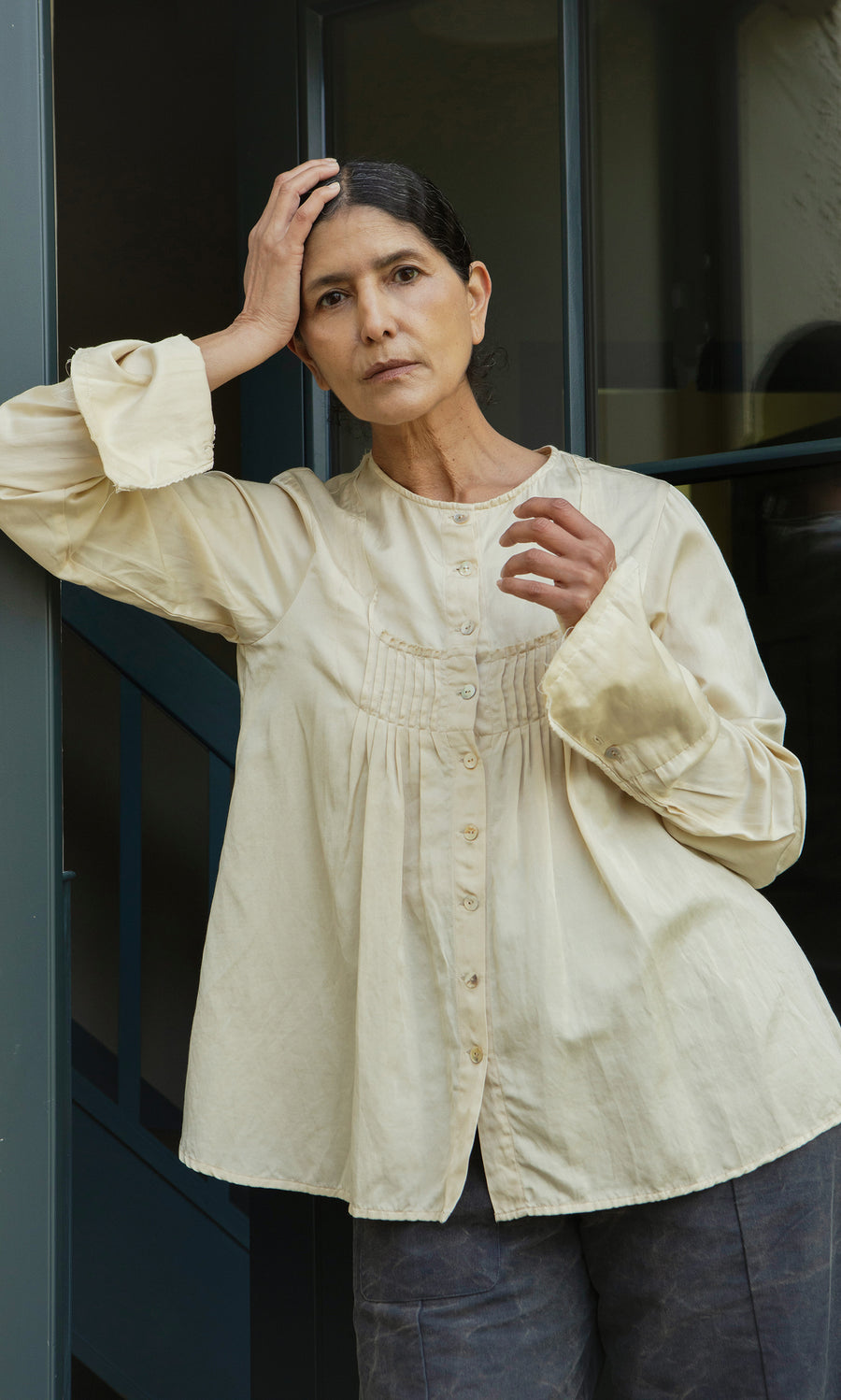 Plümo  Woman in a Silk Shirt leans in a doorway, hand on head, gazing thoughtfully at the camera.