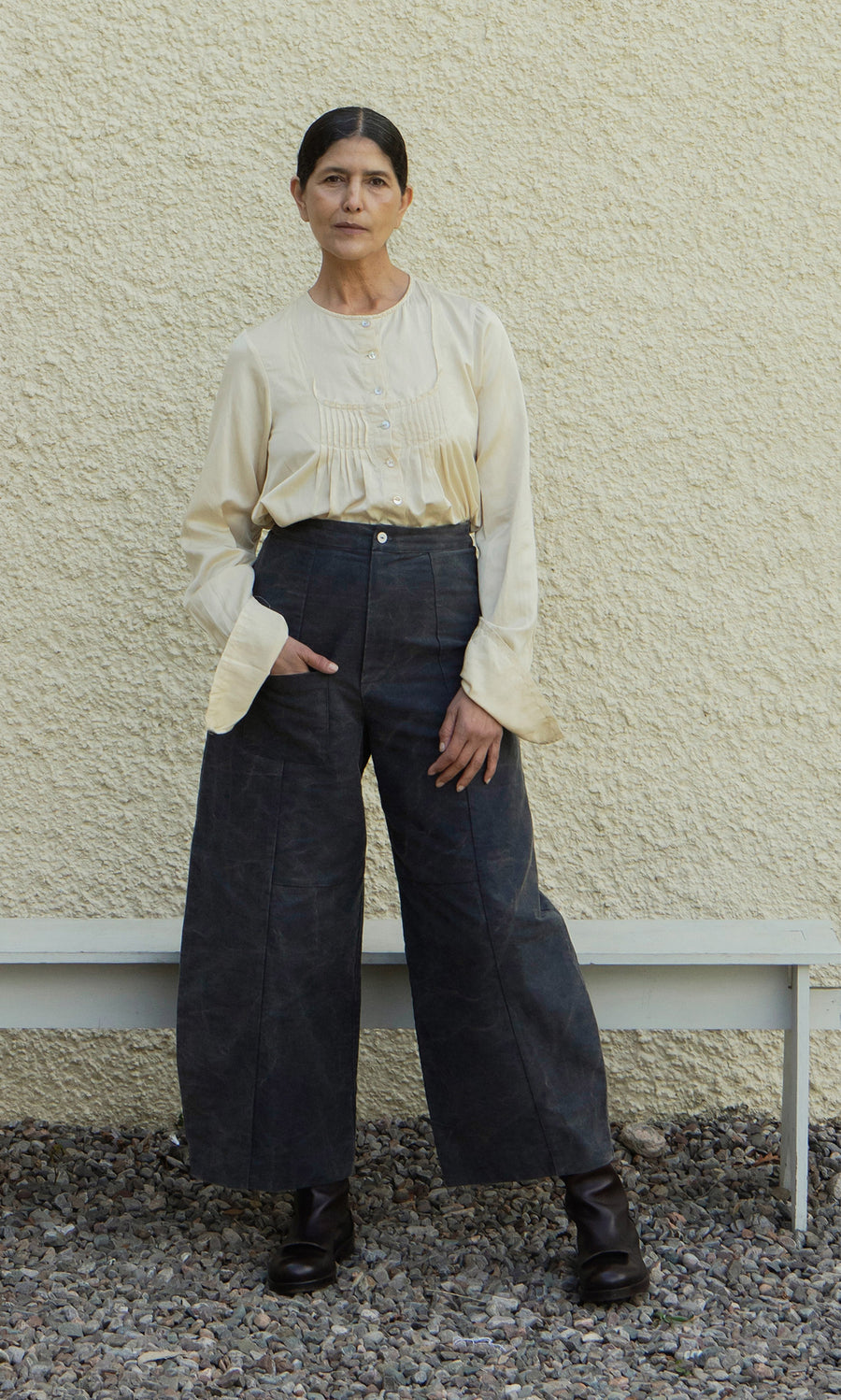 Plümo  Woman in a Silk Shirt and wide-leg dark pants stands against a textured wall, hands in pockets, looking at camera.