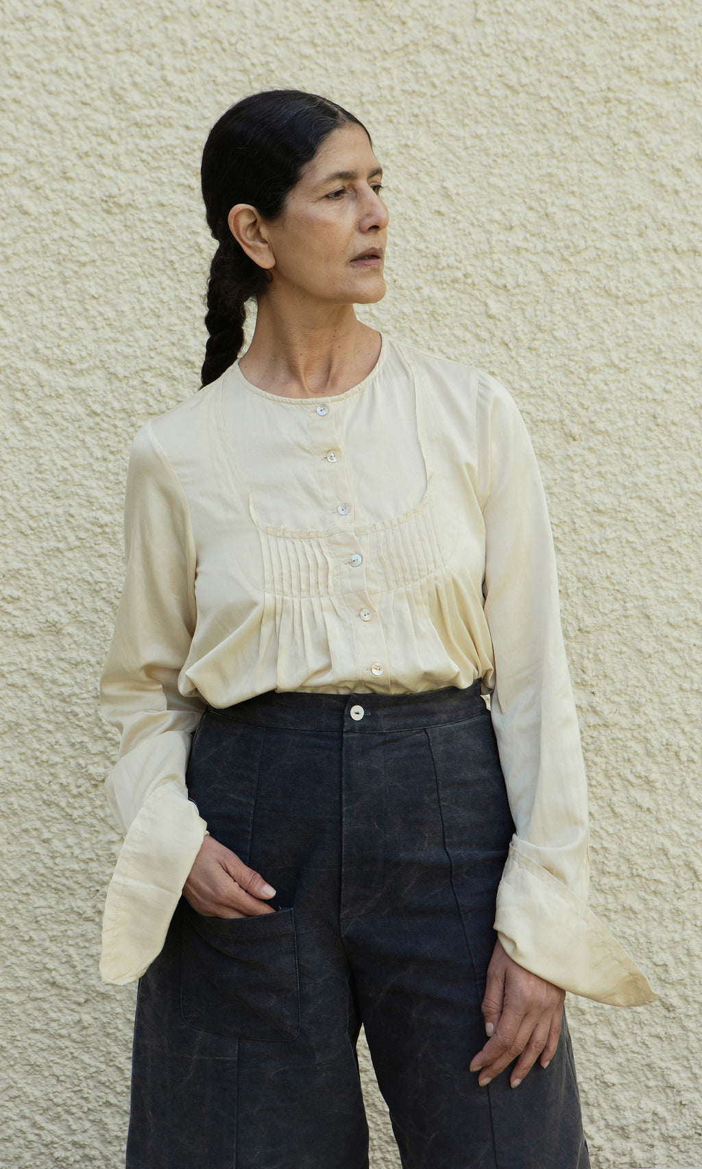 Plümo  Woman with dark braided hair in a Silk Shirt and dark pants stands against a pale wall, looking sideways.