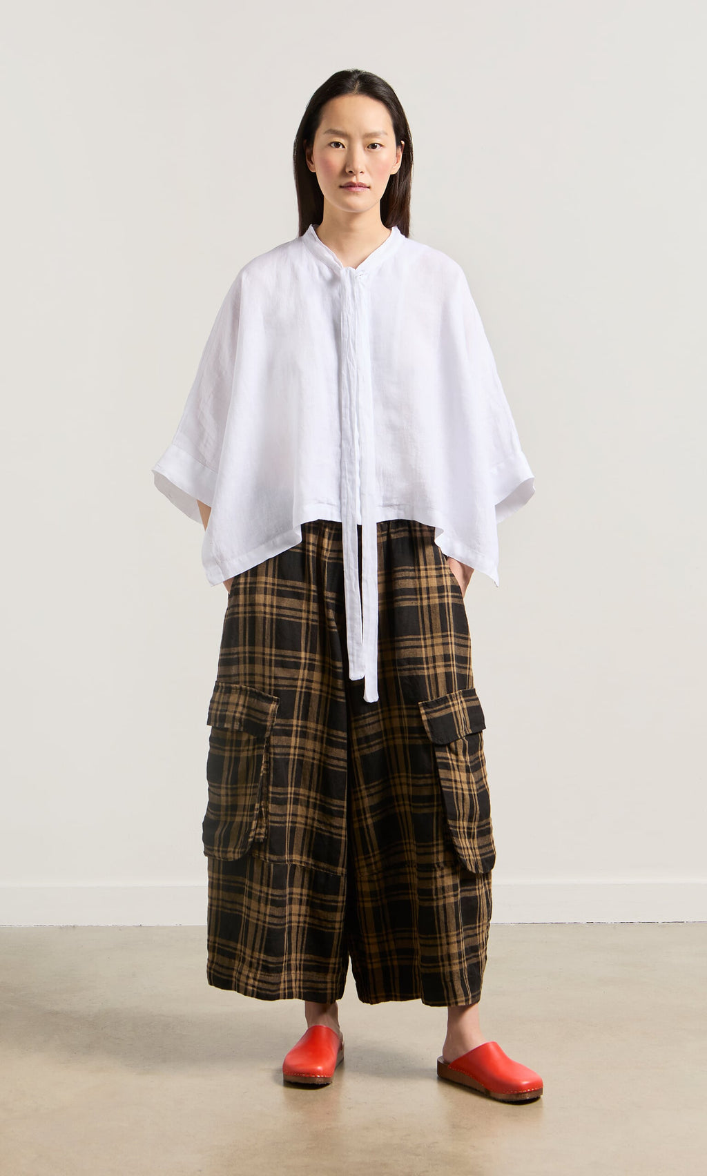 Plümo- A woman in a loose white top, Tam Check Pants with an elastic waist, and red slip-ons stands against a plain background.