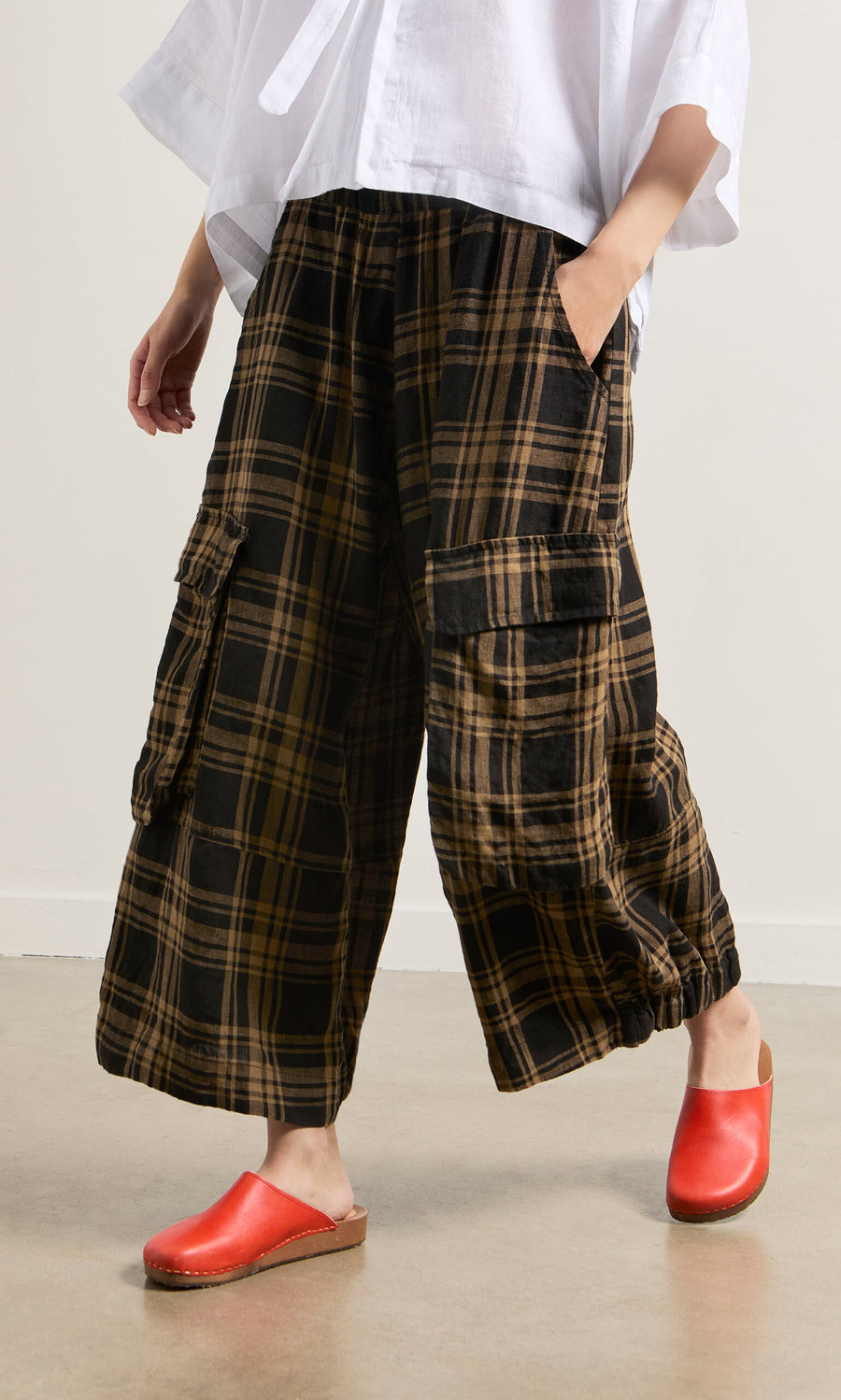 Plümo- Person wearing Tam Check Pants, a white top, and red clogs, standing indoors on a light floor.