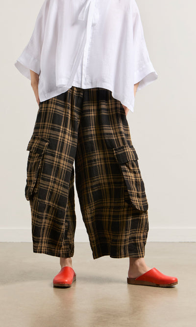 Plümo- Model in Tam Check Pants with elastic waist, loose white top, and bright red slip-ons indoors.