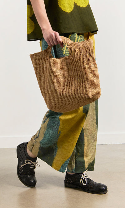 Plümo- A person holds the Tara Straw Bag while wearing patterned pants and black shoes against a plain background.