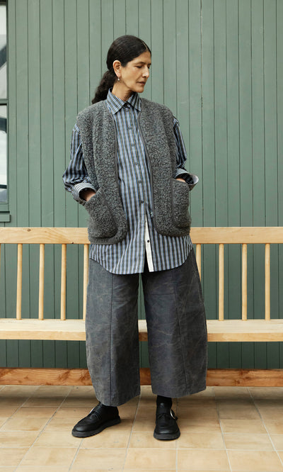 Plümo  Person wears a striped shirt, Taussig Gilet, wide pants, and black shoes before a green wall and wooden bench.