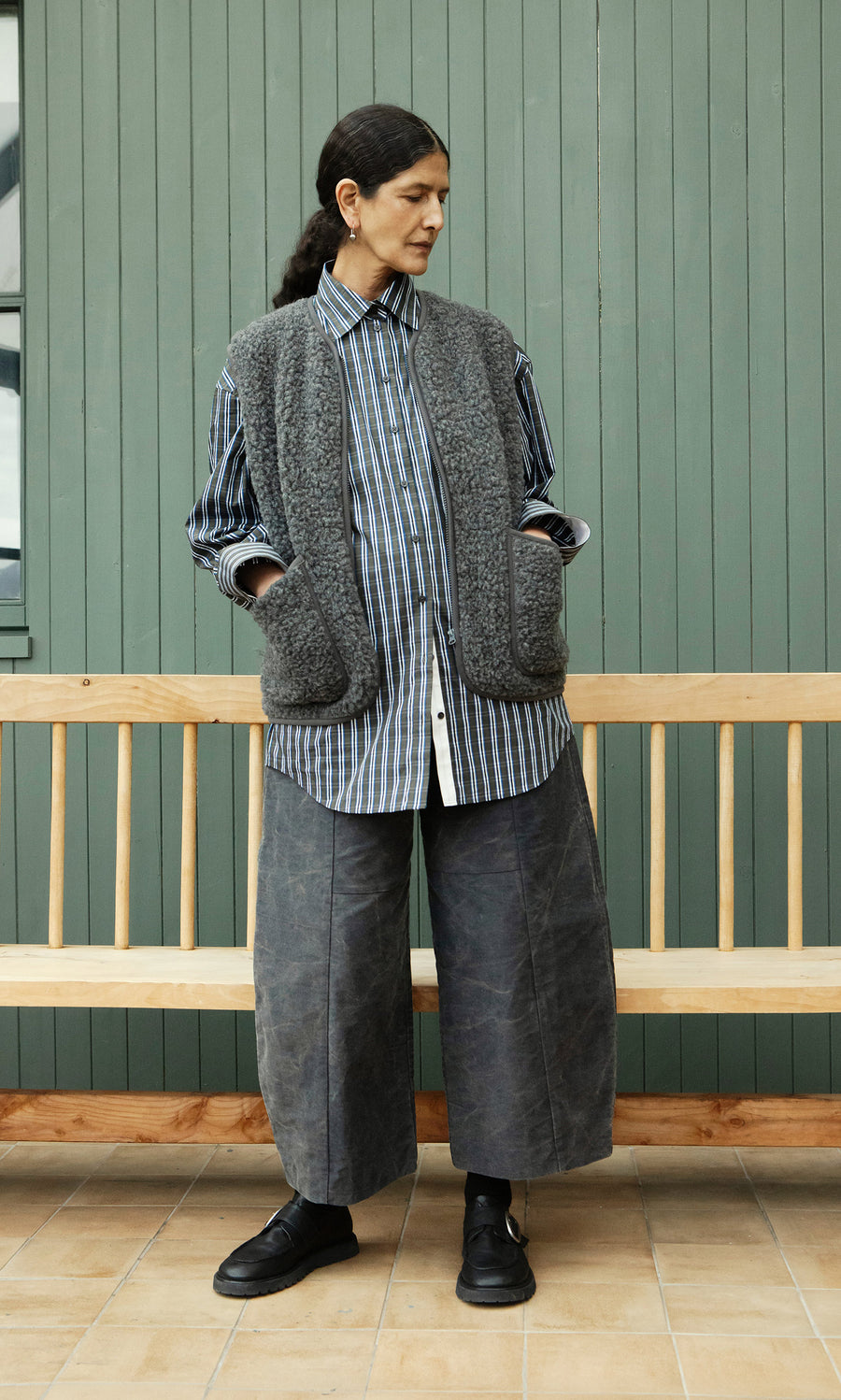 Plümo  Person wears a striped shirt, Taussig Gilet, wide pants, and black shoes before a green wall and wooden bench.