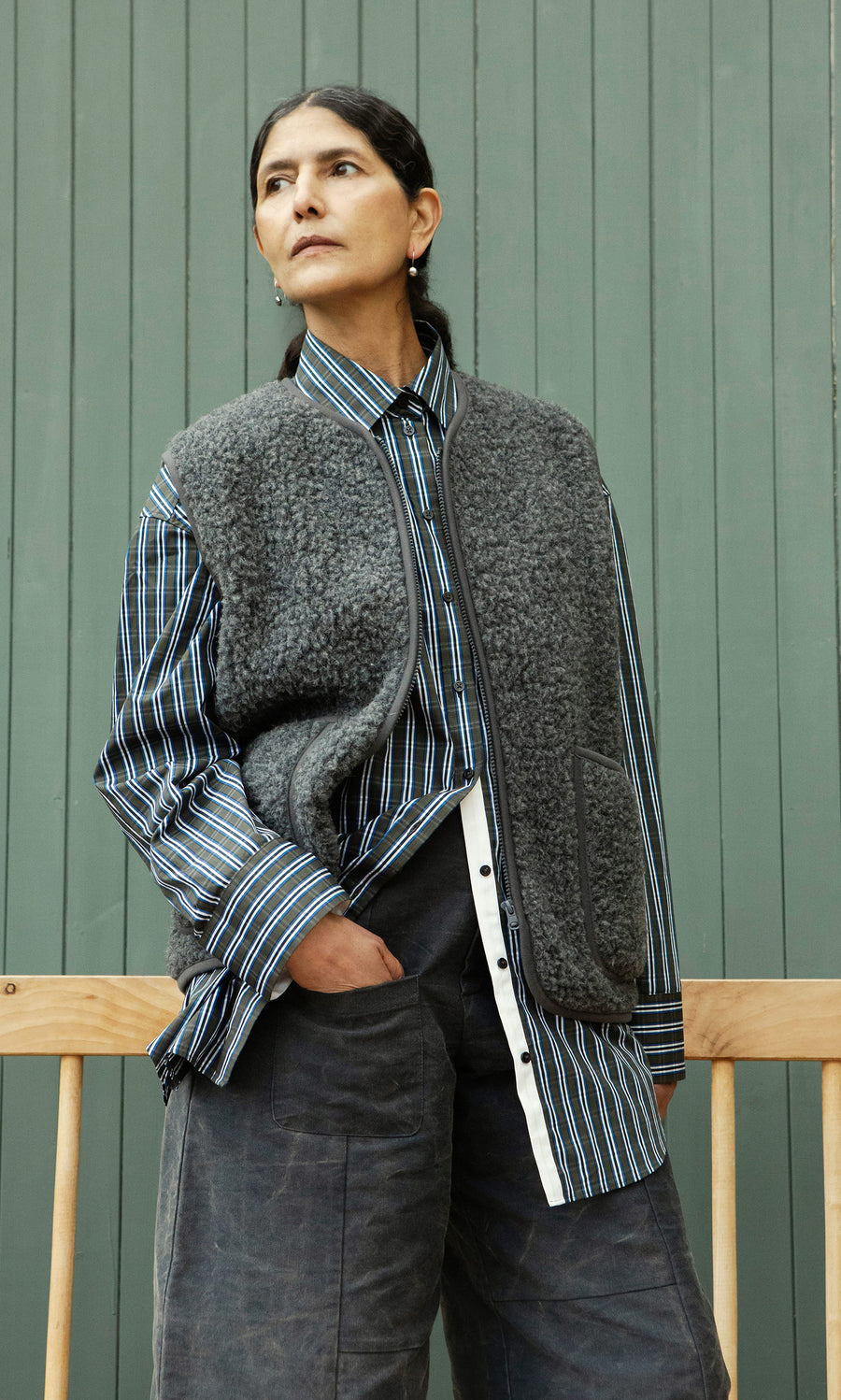 Plümo  A person in a striped shirt and the Taussig Gilet stands with one hand in their pocket, looking to the side.