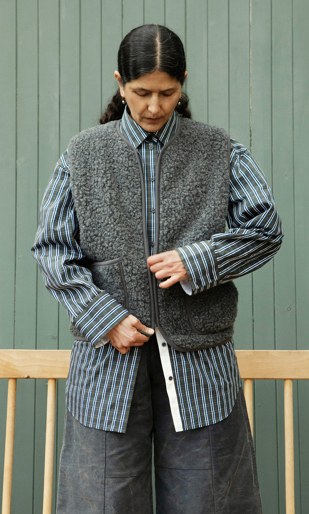 Plümo  Person in striped shirt adjusts Taussig Gilet before a green panel wall, looking down.