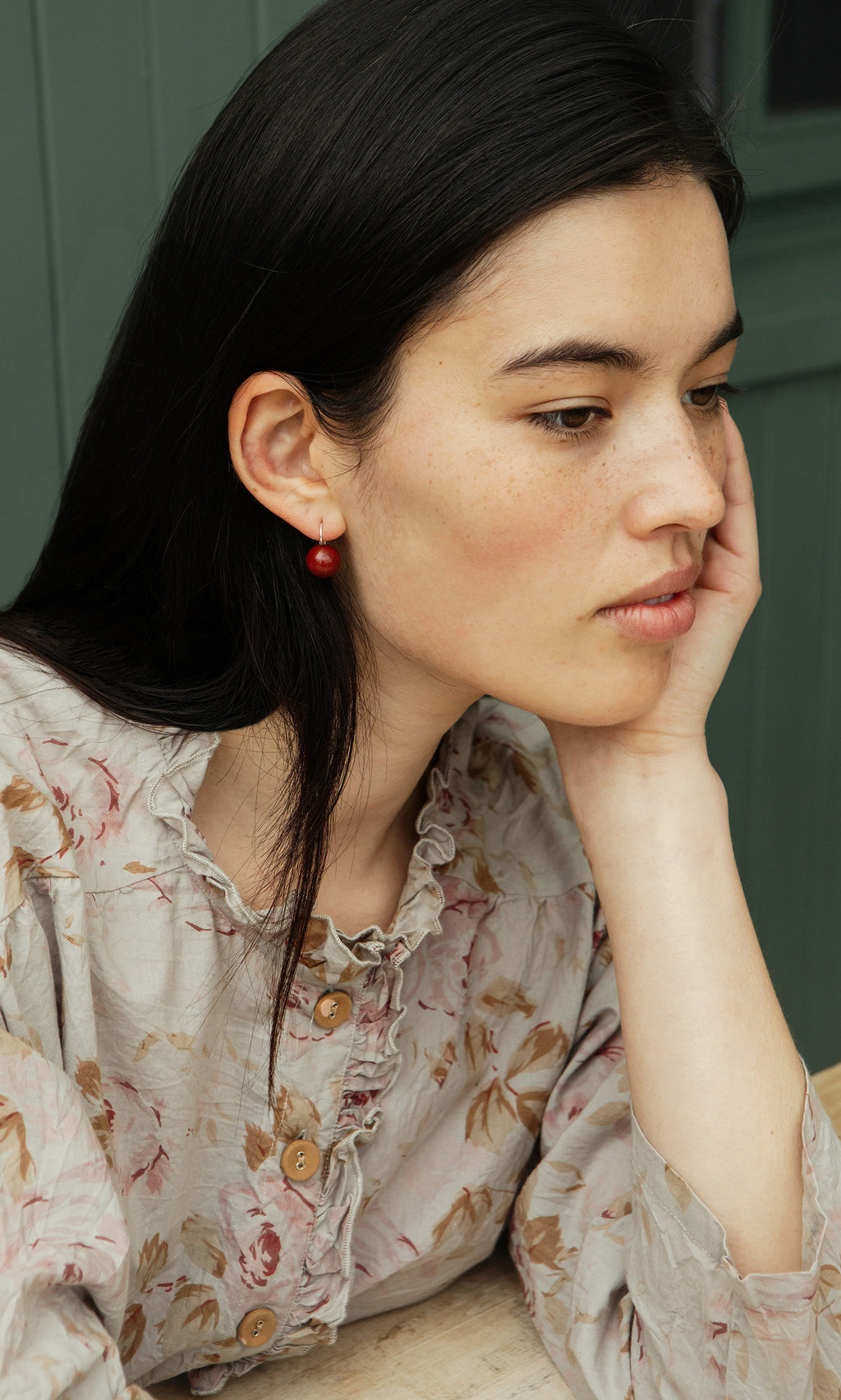 Plümo  Woman with long dark hair in a floral blouse wears Terra Earrings, resting her chin on her hand and looking thoughtful.