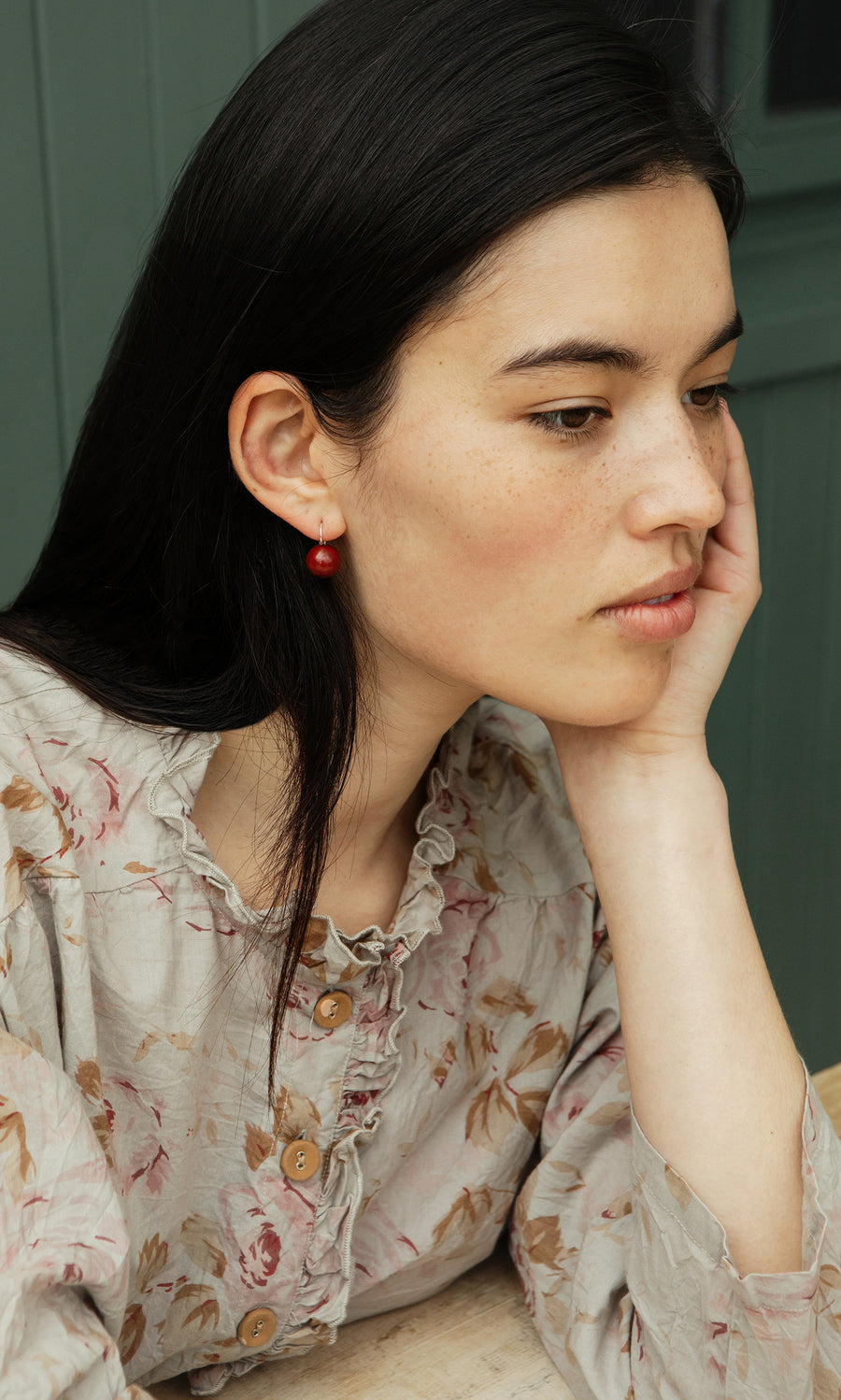 Plümo  Woman with long dark hair in a floral blouse wears Terra Earrings, resting her chin on her hand and looking thoughtful.