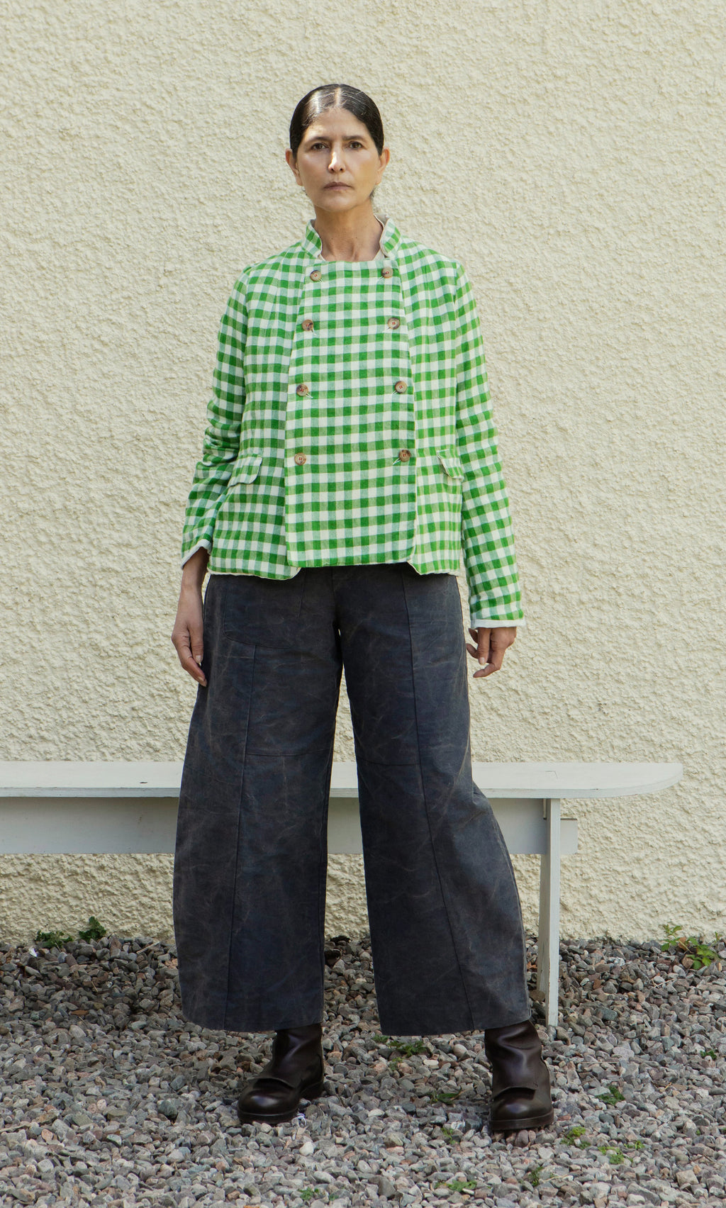 Plümo  A person in a Tiree Jacket, wide dark pants, and boots stands on gravel by a pale wall and a bench.