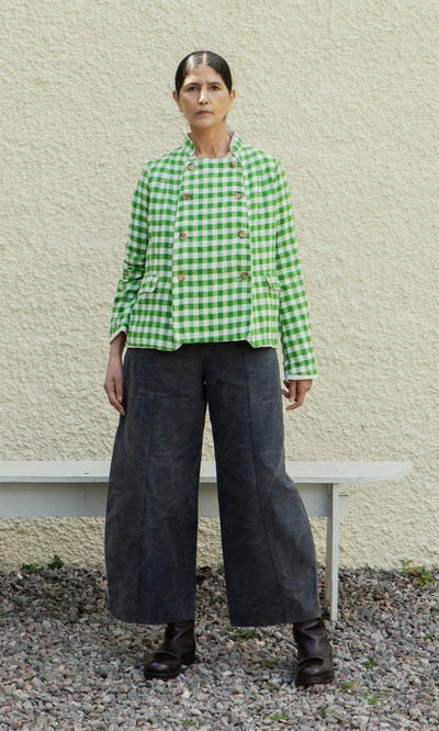 Plümo  A person in a Tiree Jacket, wide dark pants, and boots stands on gravel by a pale wall and a bench.