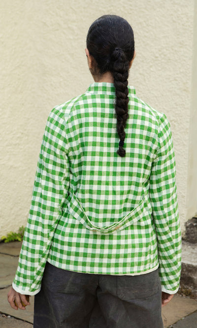 Plümo  Person in a long braid wears the Tiree Jacket, green and white gingham, facing a textured cream wall—slow fashion style.