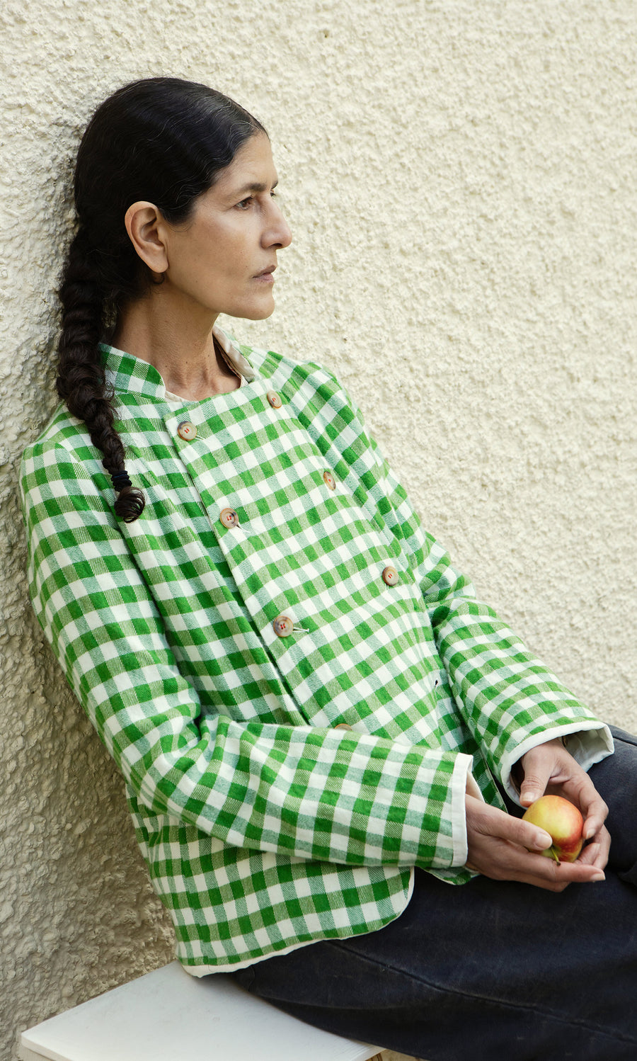Plümo  A woman with a braid in the Tiree Jacket holds an apple, seated by a textured beige wall.