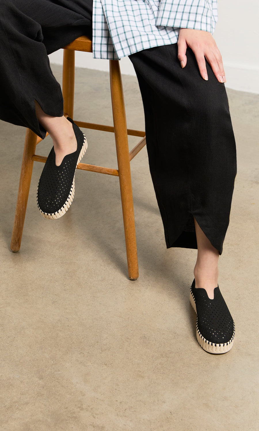 Plümo- Person sits on a wooden stool wearing Tulip Shoes - Carbon with recycled microfibre uppers on a concrete floor.
